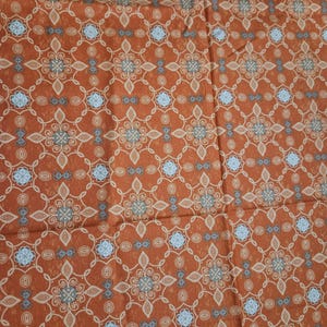 May include: Close-up of a rust-colored fabric with a repeating pattern. The design features floral and geometric shapes in light blue, gray, and beige. The fabric has a textured appearance, suggesting a woven material.