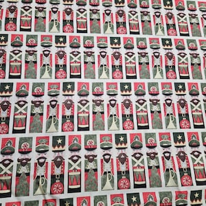 May include: White fabric with a repeating pattern of red and green Christmas nutcracker figures. The nutcrackers are holding a drum, a horn, a candy cane, and a Christmas tree ornament.