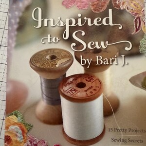 以下が含まれることがあります： Bari J. の「Inspired to Sew」というタイトルの本の表紙。木製の糸巻きが特徴です。表紙には花の装飾と「15 Pretty Projects, Sewing Secrets, Colorful Collage」というテキストが含まれています。
