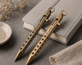 May include: Two brass-colored pens with a unique design, featuring a series of circular cutouts along the barrel. Each pen has a ring at the top and a name engraved on the side: "Dominic" and "Alexander". The pens are placed near a gray notebook.