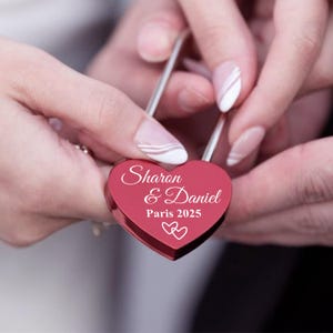 Op de afbeelding: Een rood hartvormig hangslot met de inscriptie "Sharon & Daniel Paris 2025" erop gegraveerd.