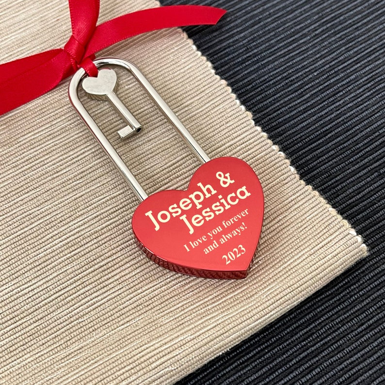 Op de afbeelding: Een zilveren hartvormig hangslot met een sleutel eraan. Het hangslot is rood en heeft de tekst "Joseph & Jessica I love you forever and always! 2023" erop gegraveerd.