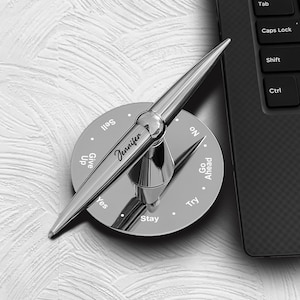 May include: A silver-colored decision maker with the name "Jennifer" engraved on it. The decision maker has a pointer and a circular base with options like "Yes", "No", "Try", and "Stay". A laptop keyboard is in the background.