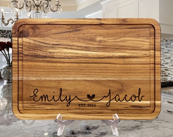 May include: A rectangular wooden cutting board with rounded corners and a juice groove. The board features a natural wood grain and is engraved with the names Emily and Jacob, a heart symbol, and the text EST. 2024 in a cursive script font.