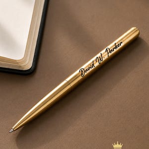 Puede incluir: Un bolígrafo de metal dorado con el nombre grabado David W. Parker en una fuente cursiva negra. El bolígrafo tiene un cuerpo cilíndrico elegante con una punta afilada y está colocado en diagonal en el encuadre junto a un cuaderno.