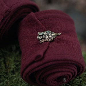 May include: A close-up of a burgundy wool shawl with a decorative metal pin in the shape of a bird. The shawl is rolled up and the pin is placed on the top of the roll.