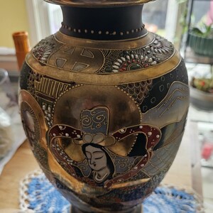May include: An ornate, vintage vase with a black neck and a gold rim. The body features detailed artwork with gold, black, and red accents, depicting a figure with a serene expression. The vase sits on a dark base.