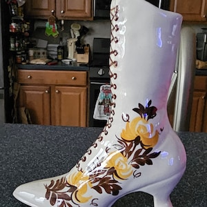 May include: White ceramic boot vase with brown and yellow floral design. The boot has a lace-up front and a high heel.