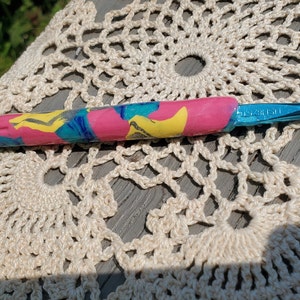 May include: A blue crochet hook with a pink, yellow, and blue patterned handle. The handle is made of polymer clay.