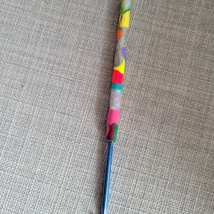 May include: A blue crochet hook with a colorful, patterned handle. The handle features sections of yellow, pink, green, and silver glitter. The hook is a crafting tool used for creating crocheted items.