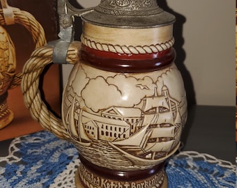 May include: A ceramic beer stein with a rope handle and a lid. The stein features a detailed illustration of a ship sailing into a harbor with buildings in the background. The text "Headed Ketsch Barkentine" is visible on the bottom of the stein.