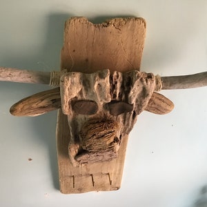 May include: A rustic, handcrafted wall hanging in the shape of a bull's head. The artwork is made from natural driftwood and features wooden horns, eyes, and a textured nose. The piece includes three small hooks at the bottom.