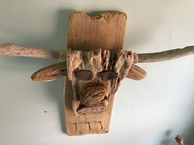 May include: A rustic, handcrafted wall hanging in the shape of a bull's head. The artwork is made from natural driftwood and features wooden horns, eyes, and a textured nose. The piece includes three small hooks at the bottom.