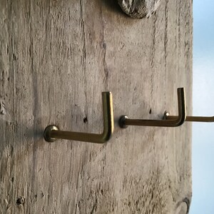 May include: A rustic wooden wall hanging with three brass hooks. The hooks are mounted on a weathered, light-colored wooden plank. The hooks are L-shaped and have a simple, minimalist design.