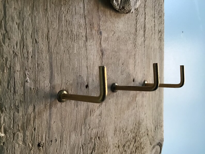 May include: A rustic wooden wall hanging with three brass hooks. The hooks are mounted on a weathered, light-colored wooden plank. The hooks are L-shaped and have a simple, minimalist design.