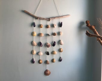 Könnte beinhalten: Ein Wandbehang bestehend aus einem horizontalen Stück Treibholz, an dem fünf vertikale Schnüre befestigt sind. Jede Schnur ist mit verschiedenen Muscheln in Weiß-, Beige-, Braun- und Violetttönen geschmückt, die kaskadenförmig angeordnet sind.