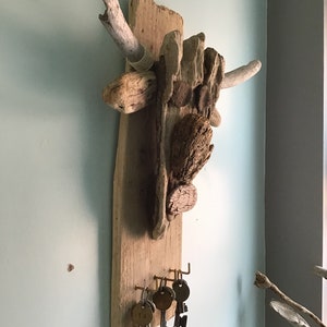 May include: A rustic key holder crafted from driftwood and various wood pieces. The wooden base features brass hooks holding several vintage keys with numbered tags. The natural wood tones and textures create a unique, organic design.