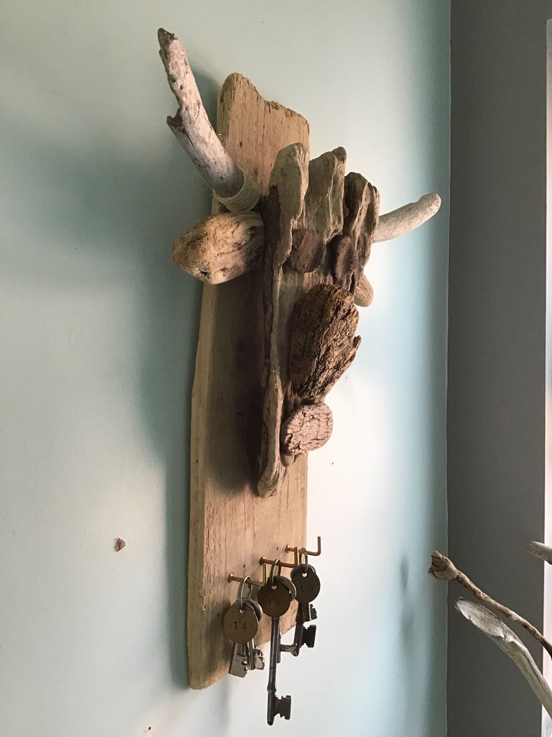 May include: A rustic key holder crafted from driftwood and various wood pieces. The wooden base features brass hooks holding several vintage keys with numbered tags. The natural wood tones and textures create a unique, organic design.