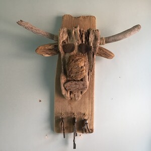 May include: A rustic wooden key holder shaped like a bull's head. The artwork features driftwood pieces, with two curved horns, and metal eye accents. Three key hooks hang below.