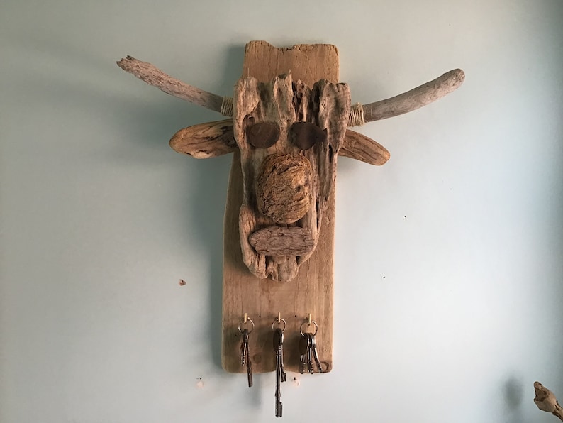 May include: A rustic wooden key holder shaped like a bull's head. The artwork features driftwood pieces, with two curved horns, and metal eye accents. Three key hooks hang below.