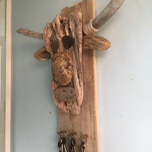 May include: A rustic driftwood key holder shaped like a bull's head, featuring natural wood tones and stone accents. The key holder has two gray wooden horns and brass hooks holding several keys. The artwork is mounted on a light blue wall.