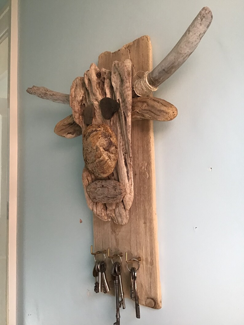 May include: A rustic driftwood key holder shaped like a bull's head, featuring natural wood tones and stone accents. The key holder has two gray wooden horns and brass hooks holding several keys. The artwork is mounted on a light blue wall.