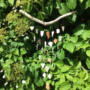 May include: A white driftwood wind chime with multiple ceramic pendants in white, brown, and orange hanging from it. The wind chime is hanging from a tree branch in a garden setting.