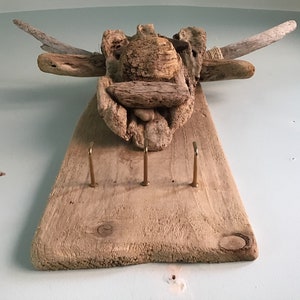 May include: A rustic, wooden key holder with three brass hooks. The key holder is made from driftwood and features a decorative top made of various pieces of wood arranged to resemble an animal head. It is mounted on a light blue wall.