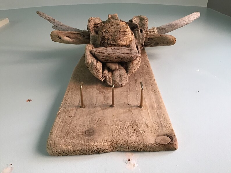 May include: A rustic, wooden key holder with three brass hooks. The key holder is made from driftwood and features a decorative top made of various pieces of wood arranged to resemble an animal head. It is mounted on a light blue wall.