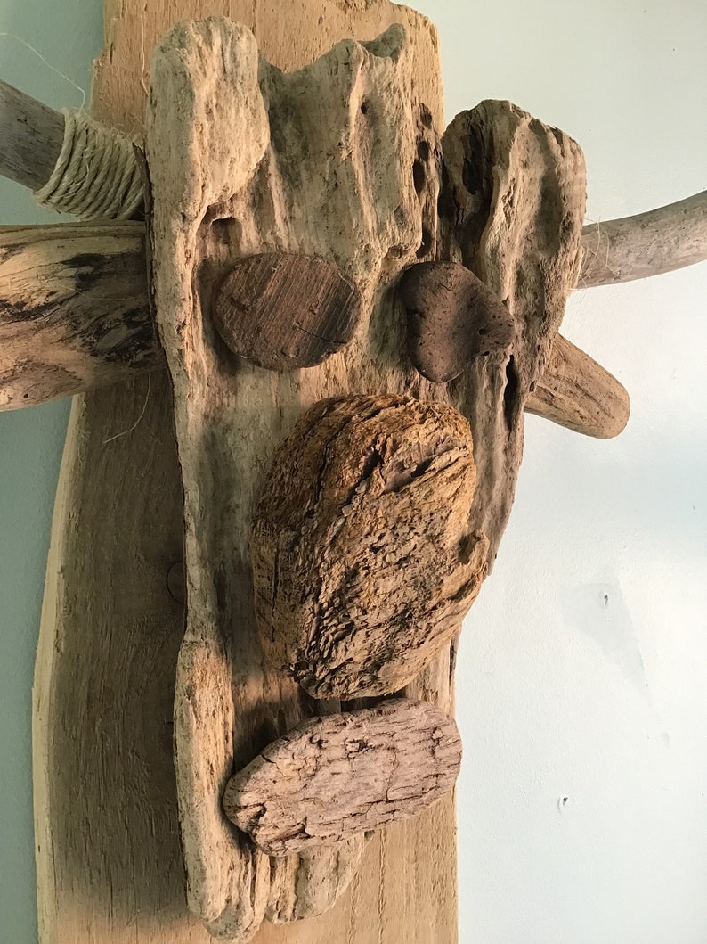 May include: A rustic wall art piece crafted from driftwood, featuring a face-like design. The artwork incorporates various wood pieces and small stones to create facial features. The color palette is primarily natural wood tones, with some rope accents.