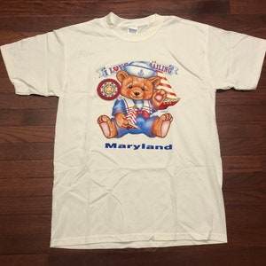 May include: White t-shirt with a cartoon bear wearing a sailor hat and holding a sailboat. The bear is surrounded by a banner that reads "I Love Sailing". The text "Maryland" is printed below the bear.