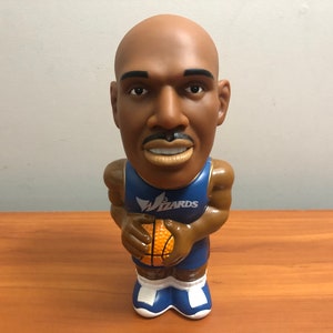 May include: A bobblehead figurine of a basketball player wearing a blue and gold Wizards jersey and holding a basketball. The player has a brown skin tone and a mustache.