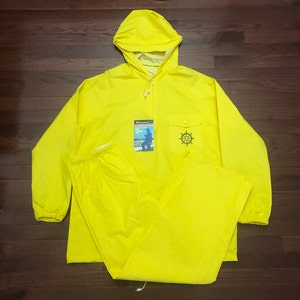 May include: A bright yellow raincoat and matching pants set with a hood. The jacket has a large pocket on the front with a nautical wheel logo. The packaging for the raincoat is visible in the image.