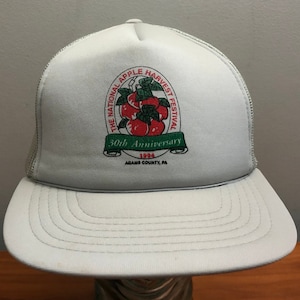 May include: White mesh trucker hat with a red, green, and white logo for the National Apple Harvest Festival. The logo features a bunch of red apples with green leaves and the text "The National Apple Harvest Festival 30th Anniversary 1994 Adams County, PA".