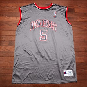 May include: A gray Champion basketball jersey with red and black trim. The jersey features the word "NEW JERSEY" arched across the chest, with the number "5" below. A small NBA logo and the Champion logo are visible.