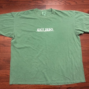 May include: A sage green t-shirt with the text "EXIT ZERO" printed in white across the chest. The shirt is laid flat on a wooden surface, showcasing its relaxed fit and casual style. The fabric appears soft and slightly faded.