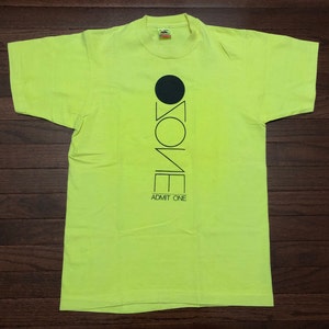 May include: Neon green t-shirt with a black graphic of the word "ZONE" with a circle above the "O". The text "ADMIT ONE" is below the graphic.