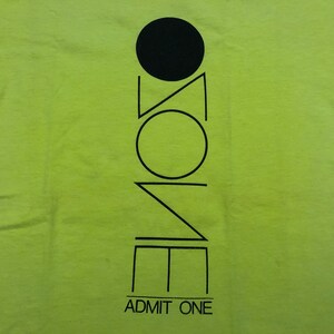 May include: A lime green t-shirt with a black graphic design of a circle, triangle, and square with the word "ZONE" written vertically inside the shapes. The text "ADMIT ONE" is printed below the graphic.