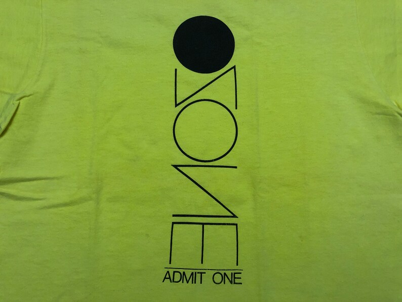 May include: A lime green t-shirt with a black graphic design of a circle, triangle, and square with the word "ZONE" written vertically inside the shapes. The text "ADMIT ONE" is printed below the graphic.