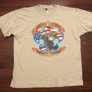 May include: A beige t-shirt with a graphic of a bald eagle and a bluebird in front of an American flag. The graphic is surrounded by a gold fleur-de-lis and the text "Camp Lions 2002 Wild Scout Reservation Tidewater Council B.S.A."