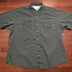 May include: A short-sleeved button-down shirt in a dark gray color with a repeating geometric pattern. The shirt has two chest pockets with button closures and a classic collar. The shirt is laid flat on a wooden surface.