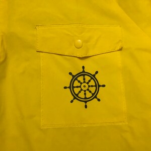 May include: A yellow raincoat with a black steering wheel logo on the front pocket. The pocket has a yellow button.