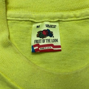 May include: A yellow Fruit of the Loom t-shirt with a tag that says "100% Cotton Preshrunk" and "Fruit of the Loom" with a logo of a bunch of fruit. The tag also says "Made in U.S.A." and "M" for size.