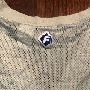 May include: White mesh jersey with a blue diamond-shaped tag with the letter "F" in the center and the trademark symbol.