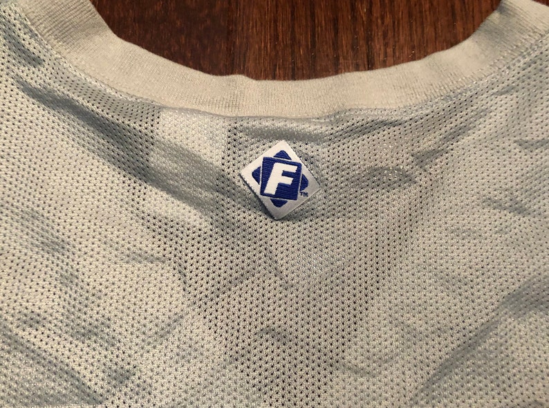 May include: White mesh jersey with a blue diamond-shaped tag with the letter "F" in the center and the trademark symbol.