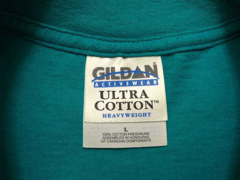 May include: A white tag with black text that reads "GILDAN ACTIVEWEAR ULTRA COTTON&trade; HEAVYWEIGHT" and "100% COTTON PRESHRUNK ASSEMBLED IN HONDURAS OF CANADIAN COMPONENTS". The tag is attached to a teal shirt.