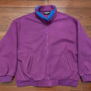 May include: A purple fleece zip-up jacket with a blue inner collar and a zipper closure. The jacket has two pockets on the front and a ribbed waistband.