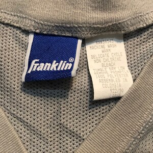 May include: A blue care label with the text "Franklin" and care instructions for the garment. The care instructions include machine wash warm, delicate cycle, non chlorine bleach, tumble dry low, remove promptly, 100% polyester, assembled in Colombia, RN#59543, 922/90.