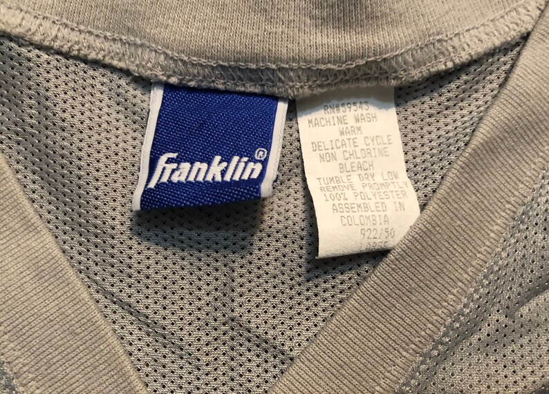 May include: A blue care label with the text "Franklin" and care instructions for the garment. The care instructions include machine wash warm, delicate cycle, non chlorine bleach, tumble dry low, remove promptly, 100% polyester, assembled in Colombia, RN#59543, 922/90.