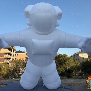 Op de afbeelding: Een grote, witte opblaasbare astronautfiguur met uitgestrekte armen en een knielende houding. De figuur heeft een helm en een rechthoekig borststuk. De achtergrond toont gebouwen en een blauwe lucht.
